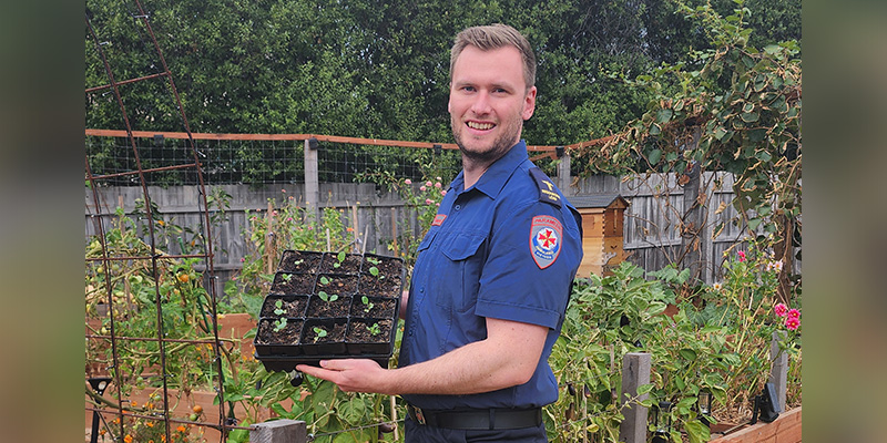 AV6K paramedic Ash Burke is standing in his garden and holding a container with several small pots and fresh saplings growing out of the soil.