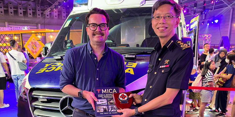 AV6K Medical Director Associate Professor David Anderson presenting the Mobile Intensive Care Ambulance (MICA) book to SCDF Director of Emergency Medical Services Department Senior Assistant Commissioner Yong Meng Wah in Singapore in 2023. Both men were standing in front of an ambulance in an exhibition hall.